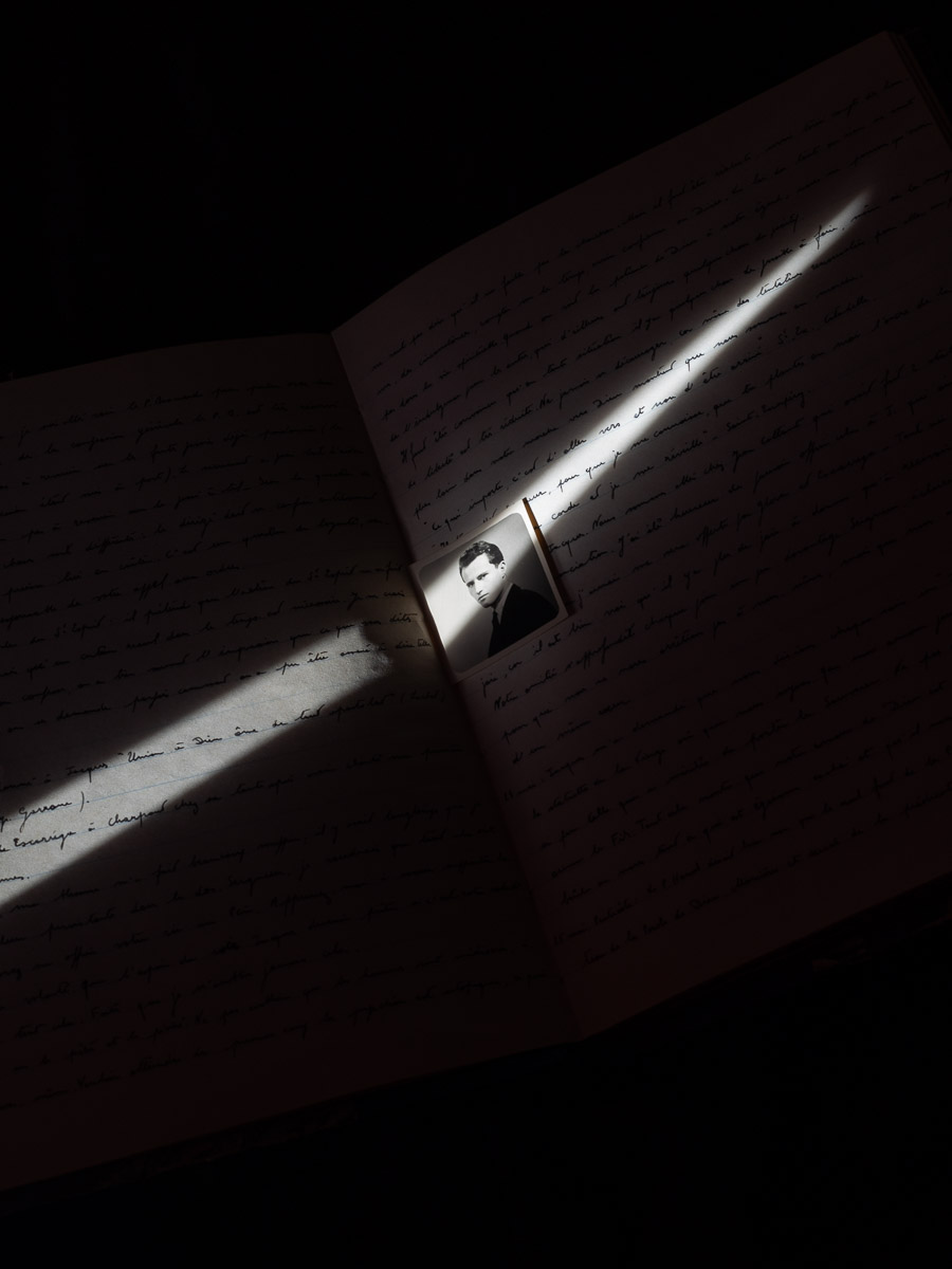 Un carnet éclairé partiellement par un rai de lumière, on aperçoit une écriture manuscrite et la photographie d'un homme glissée entre les pages.